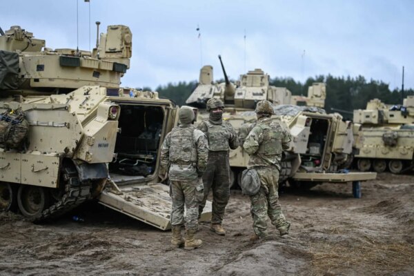 Soldiers in camouflage near the open rear of a pale armored vehicle standing on mud.