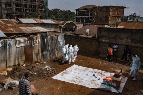 An open dirt area with buildings and corrugated metal structures. Several people in white protective suits are gathered, while another person in a light blue suit and apron stands near a white tarp and box.