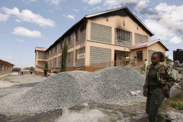 A building with beige walls and gray blocks has visible burn damage on its upper floor. A person in camouflage uniform walks in the foreground, with a large pile of gray gravel nearby.