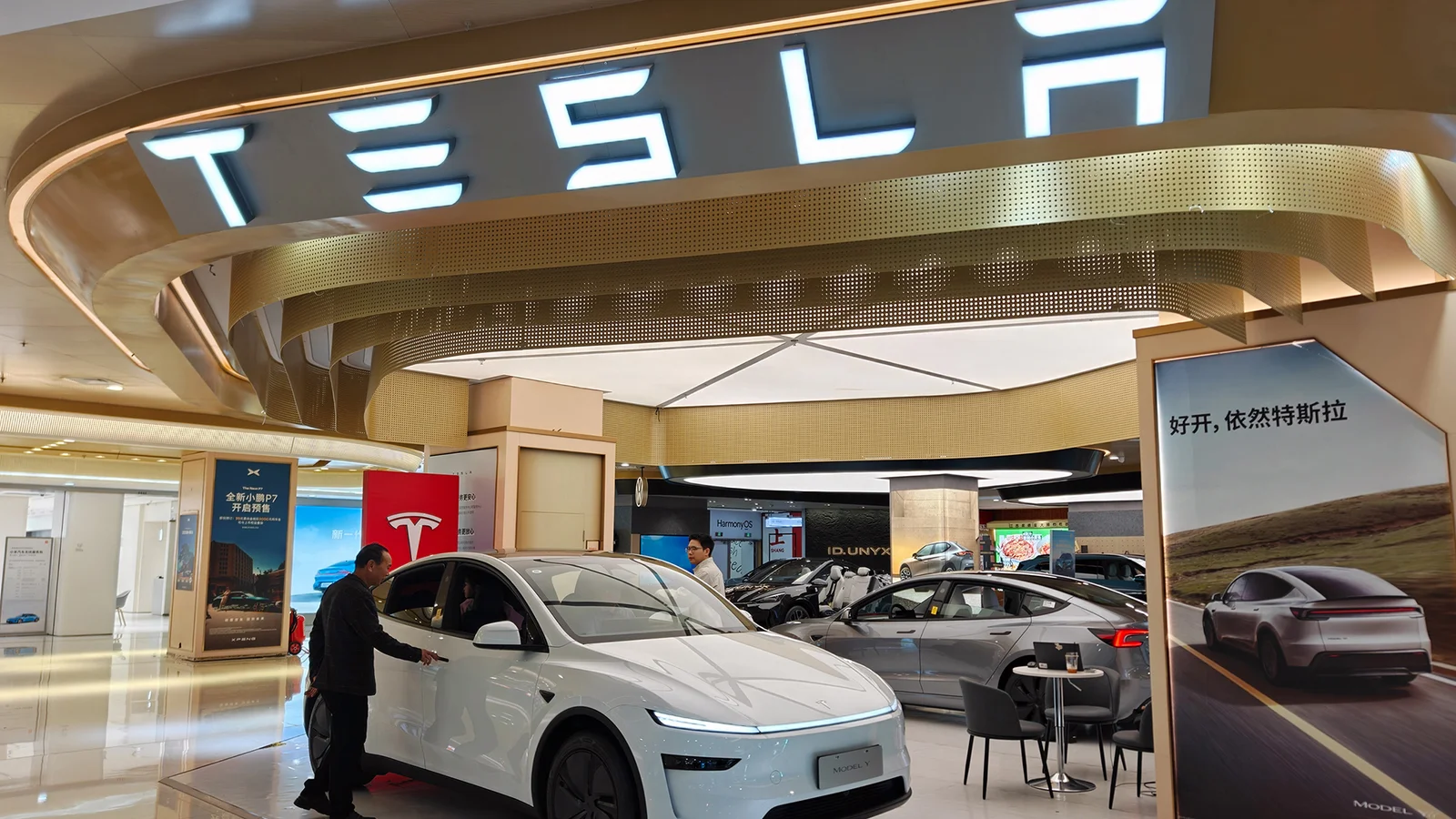 A white Tesla vehicle displayed in a modern showroom, with a man inspecting it under prominent Tesla signage.