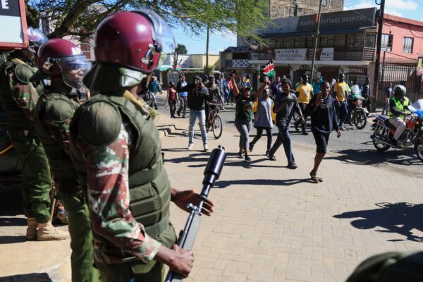 Three people in military uniforms and maroon helmets, one with a black gun. A crowd walks on the street, some raising arms and carrying flags.