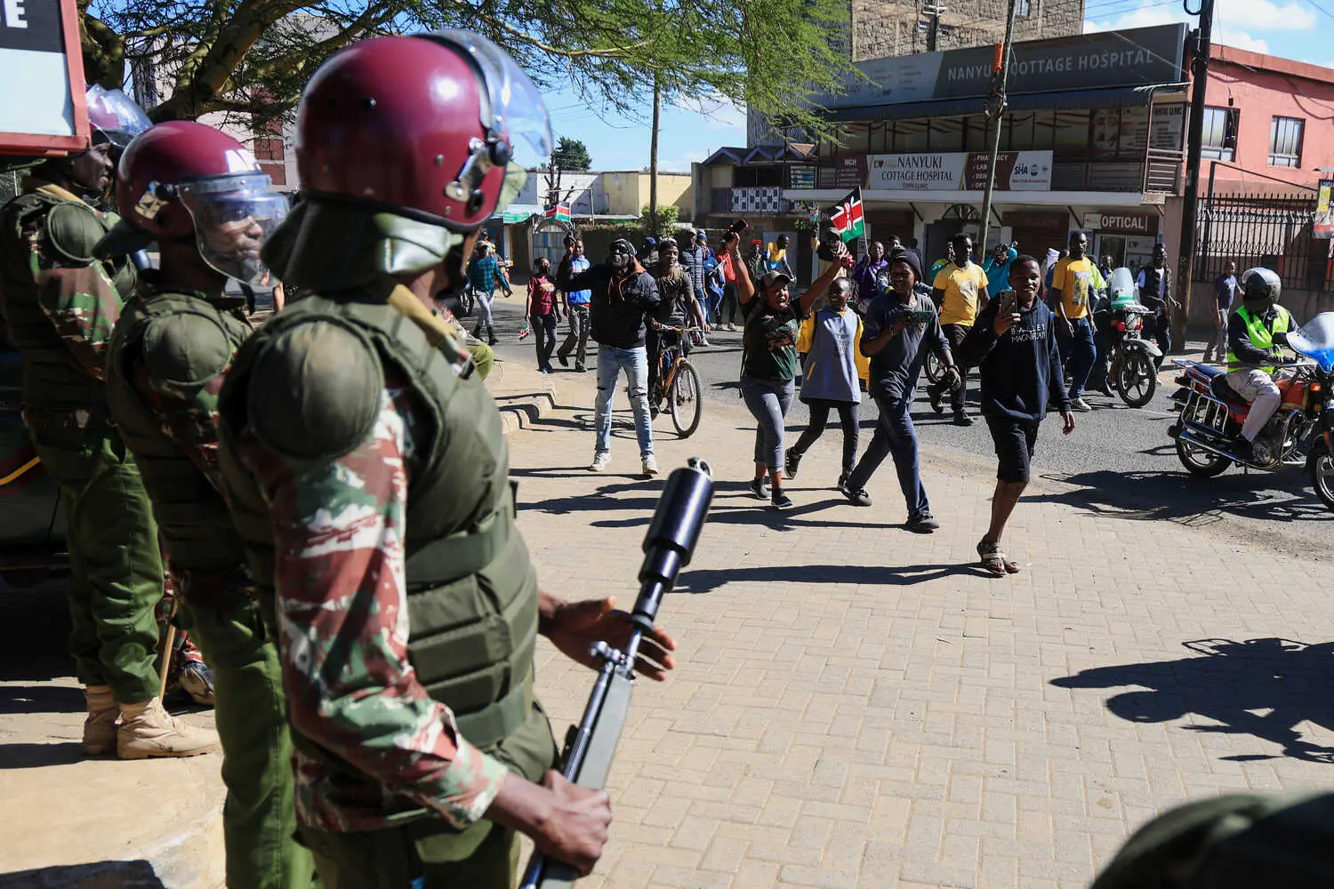Three people in military uniforms and maroon helmets, one with a black gun. A crowd walks on the street, some raising arms and carrying flags.