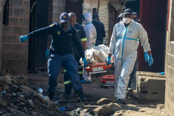 Several people, some in police clothing and others in white protective gear, carry a stretcher with a body covered by a sheet. They walk on a path covered with debris.