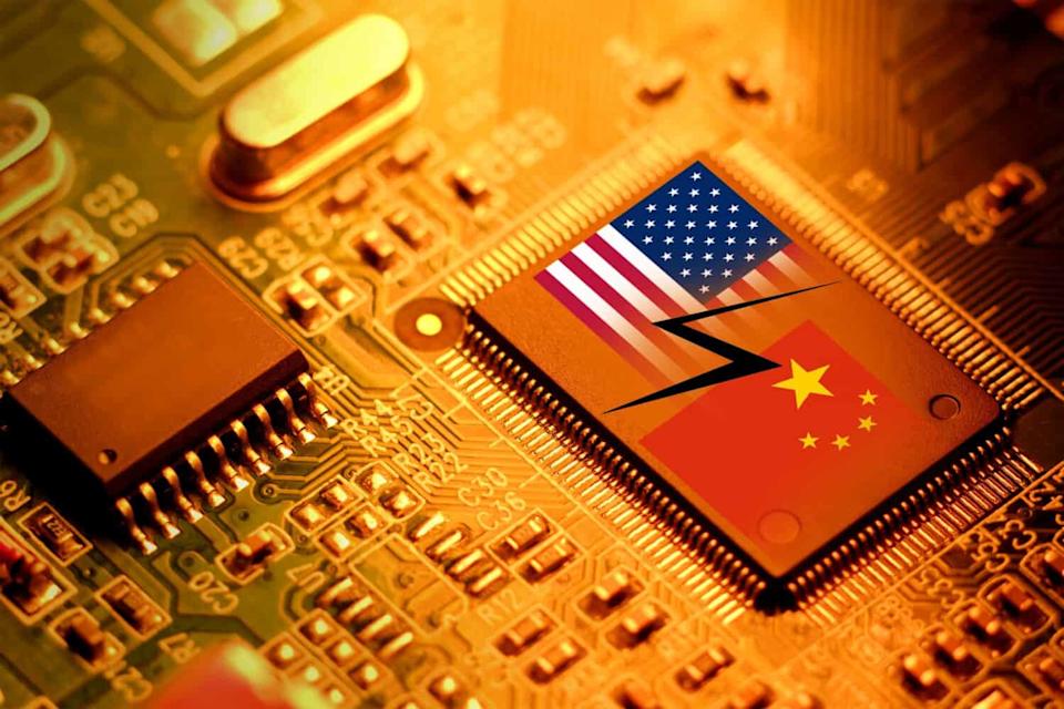 A close-up photograph of a golden-orange circuit board with various electronic components. In the foreground, a prominent rectangular microchip displays the American flag on its left half and the Chinese flag on its right half, separated by a jagged black lightning bolt. The circuit board and components are bathed in warm, glowing light.