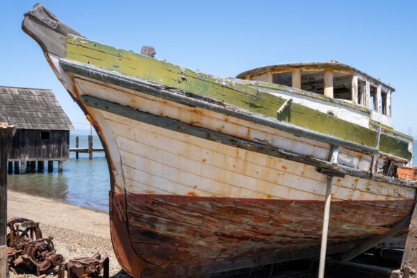 An abandoned wooden boat with peeling paint and rust sits on a shore near a pier and a weathered shed.