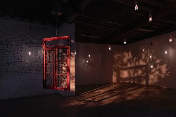 A dim installation space shows a red-framed window set into a brick wall, with small hanging lights casting shadow patterns across the floor and walls.