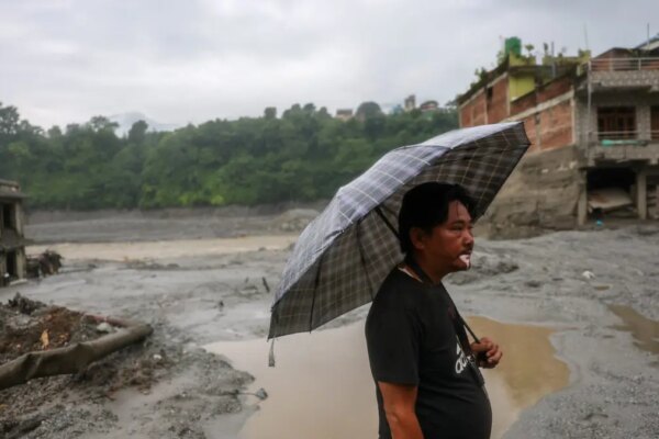 Man holds an umbrella standing before damaged buildings in Trishuli, Nepal.