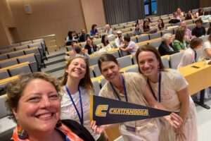 Four attendees smile for a selfie in a lecture hall during an academic conference. Dr. Ashleigh Pipes, second from the right, holds a black-and-gold University of Tennessee at Chattanooga pennant while standing with three colleagues. Other conference participants are seated and talking in the background.