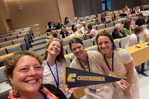 Four attendees smile for a selfie in a lecture hall during an academic conference. Dr. Ashleigh Pipes, second from the right, holds a black-and-gold University of Tennessee at Chattanooga pennant while standing with three colleagues. Other conference participants are seated and talking in the background.