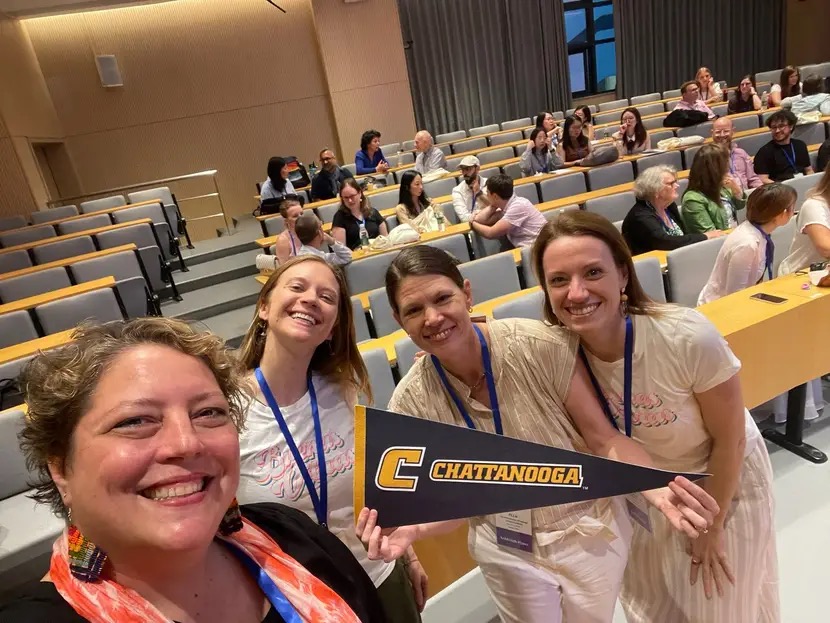 Four attendees smile for a selfie in a lecture hall during an academic conference. Dr. Ashleigh Pipes, second from the right, holds a black-and-gold University of Tennessee at Chattanooga pennant while standing with three colleagues. Other conference participants are seated and talking in the background.