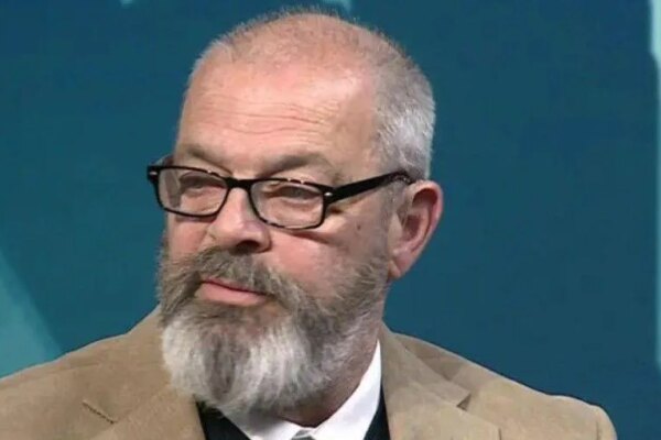 A man with a grey and brown beard and short grey hair which is mainly on the sides of his head. He is wearing glasses with a dark frame. He is wearing a beige suit jacket and a white shirt.
