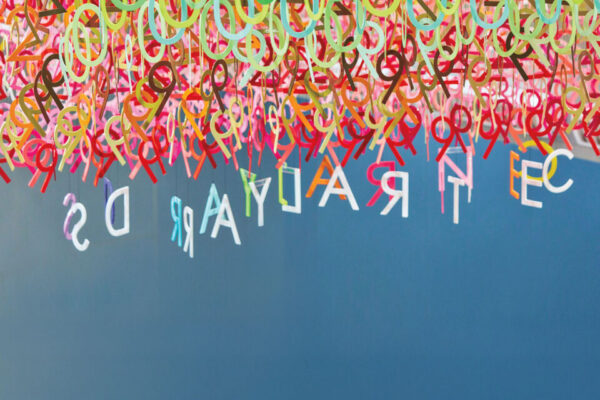 Colorful plastic letters hang densely from the ceiling, with a few letters spaced out against a blue background, appearing upside down.