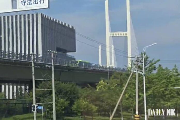 A green bus travels along an elevated approach road leading to the New Yalu River Bridge in Dandong, China, with the bridge's white cable-stayed tower visible in the background.